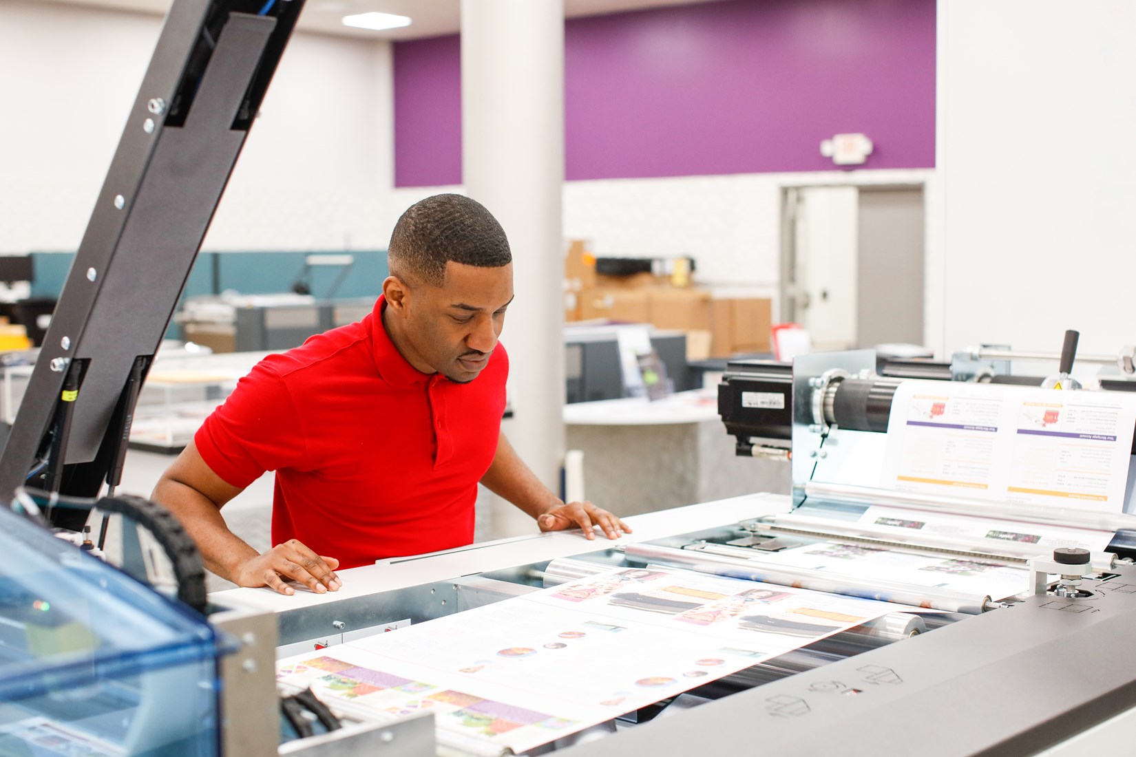 A man in a red shirt carefully examines printed materials coming out of a large industrial printer in a modern print shop.